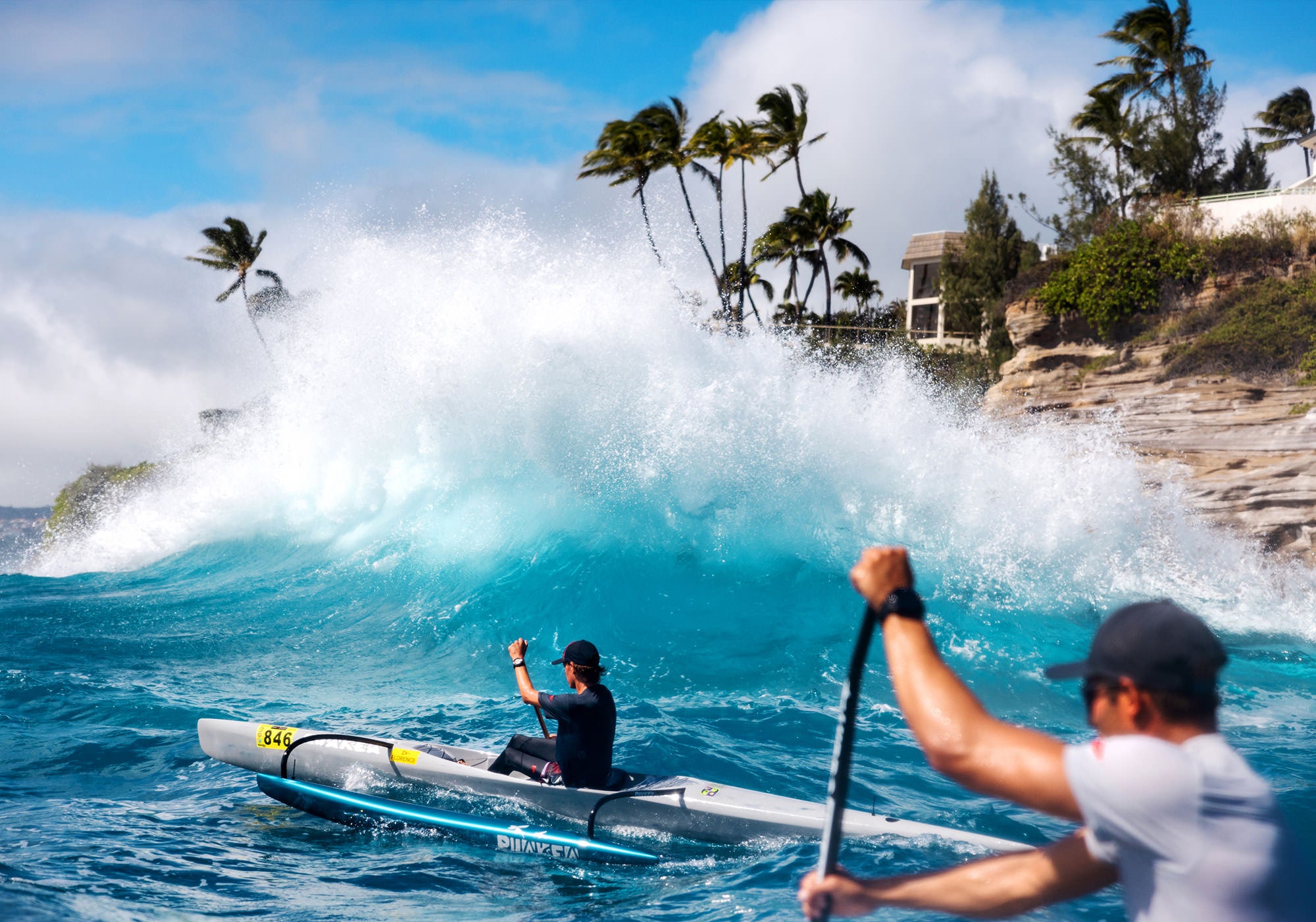 Two people outrigger paddling in separate canoes in the ocean with a wave crashing on the rocks in a tropical setting