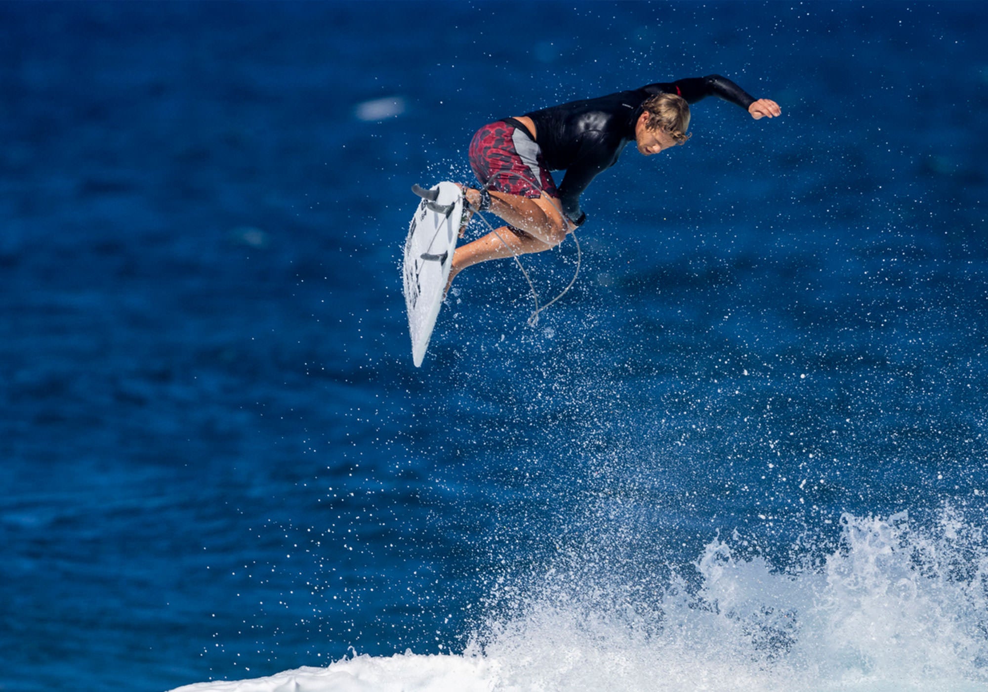 John John Florence in rotation in the air while completing an air trick while surfing, wearing a rashguard and boardshorts