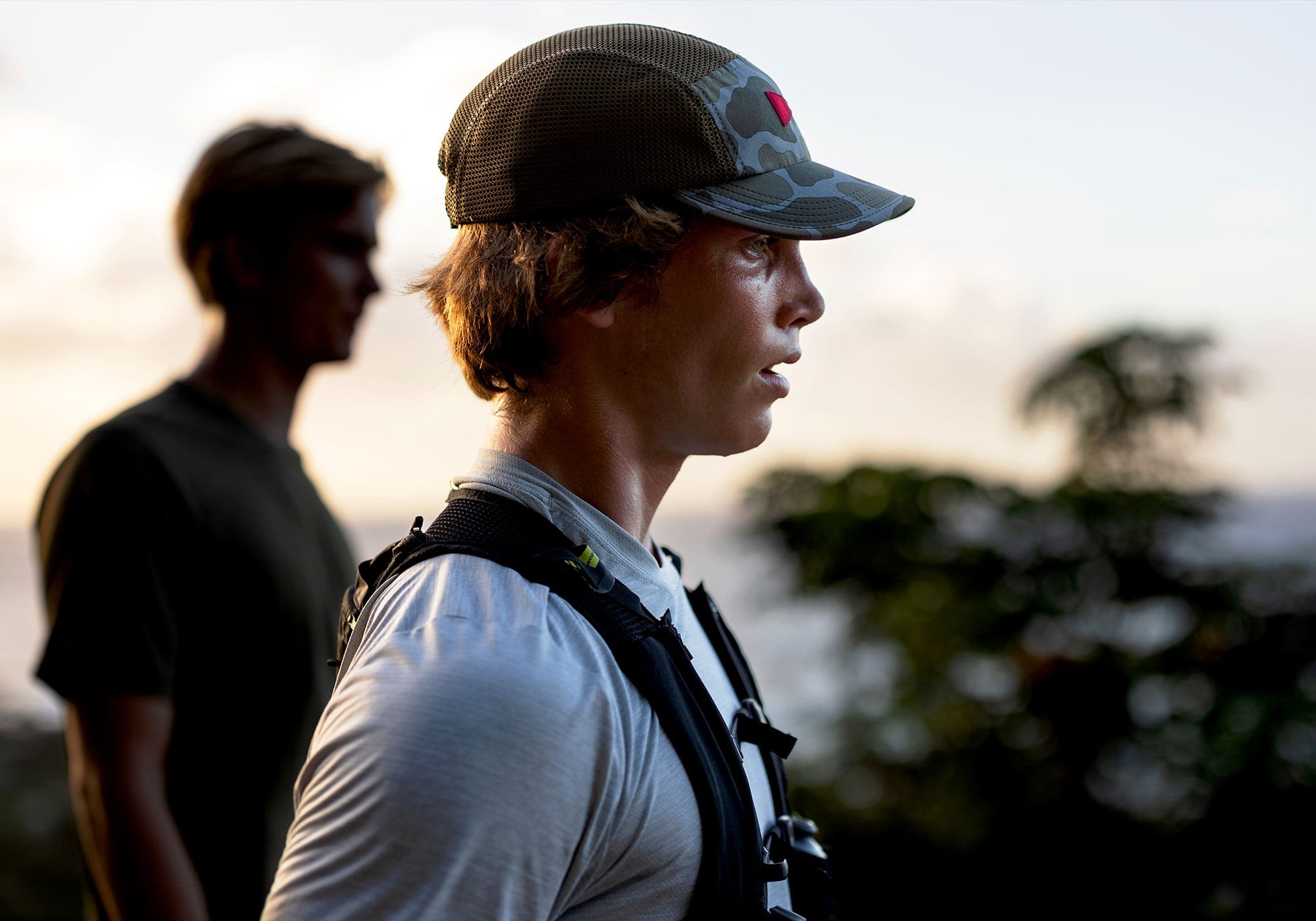Side profile of a male athlete in the foreground, wearing a camouflage Florence Hat, who appears to have just finished a hike, with a second male athlete blurred in the background