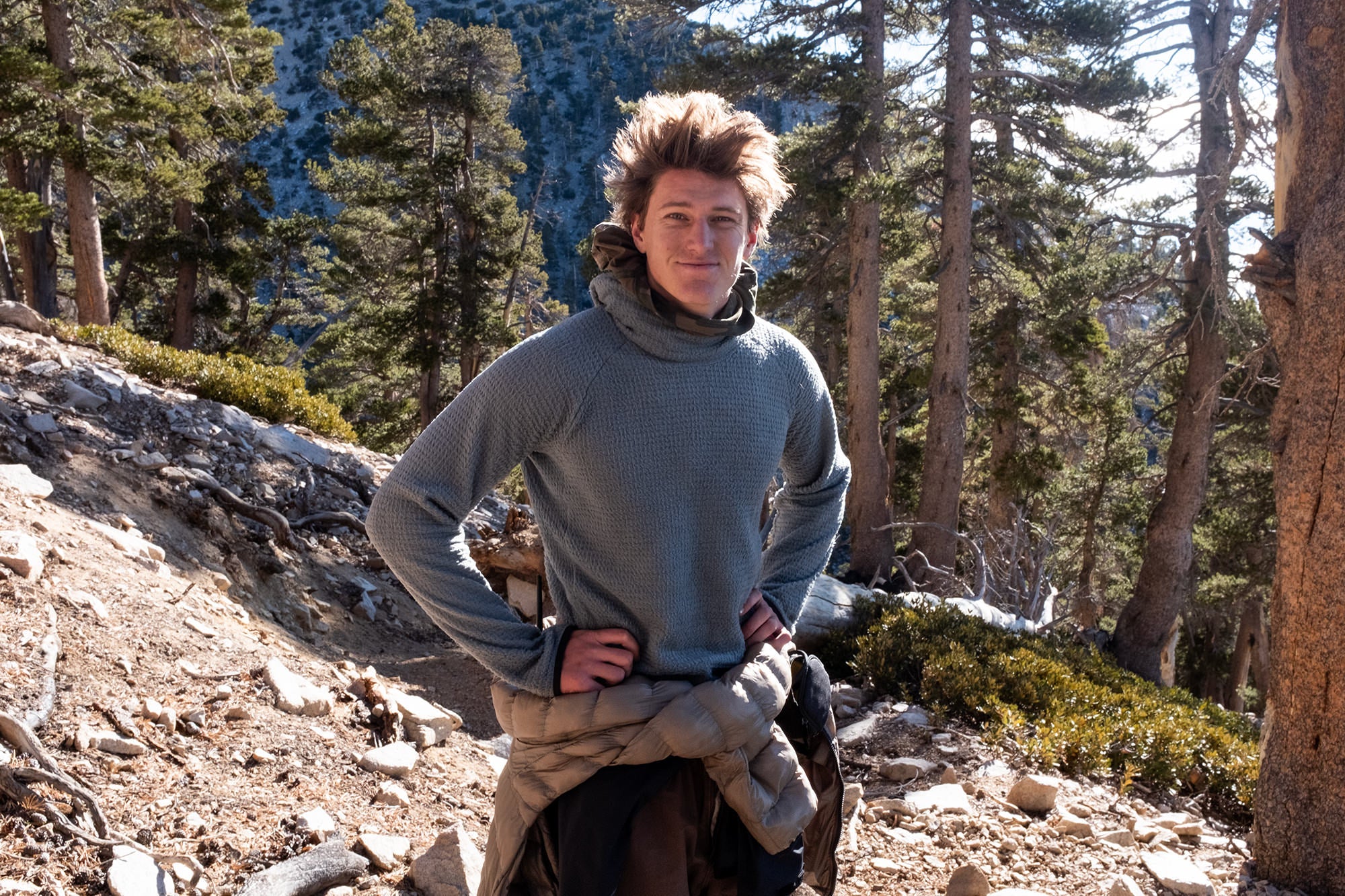 Man in the trees in the mountains wearing multiple layers of Florence jackets and fleece, while looking into the camera