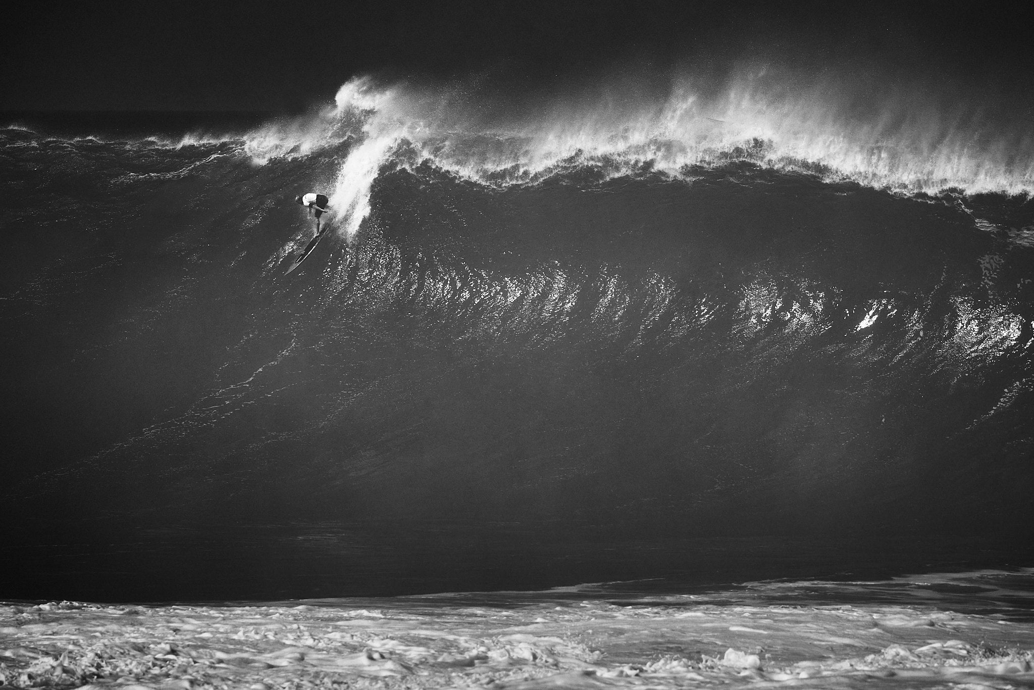 Black and white image of John John Florence dropping into a large wave on his surfboard at Waimea Bay during the Eddie Aikau memorial contest 