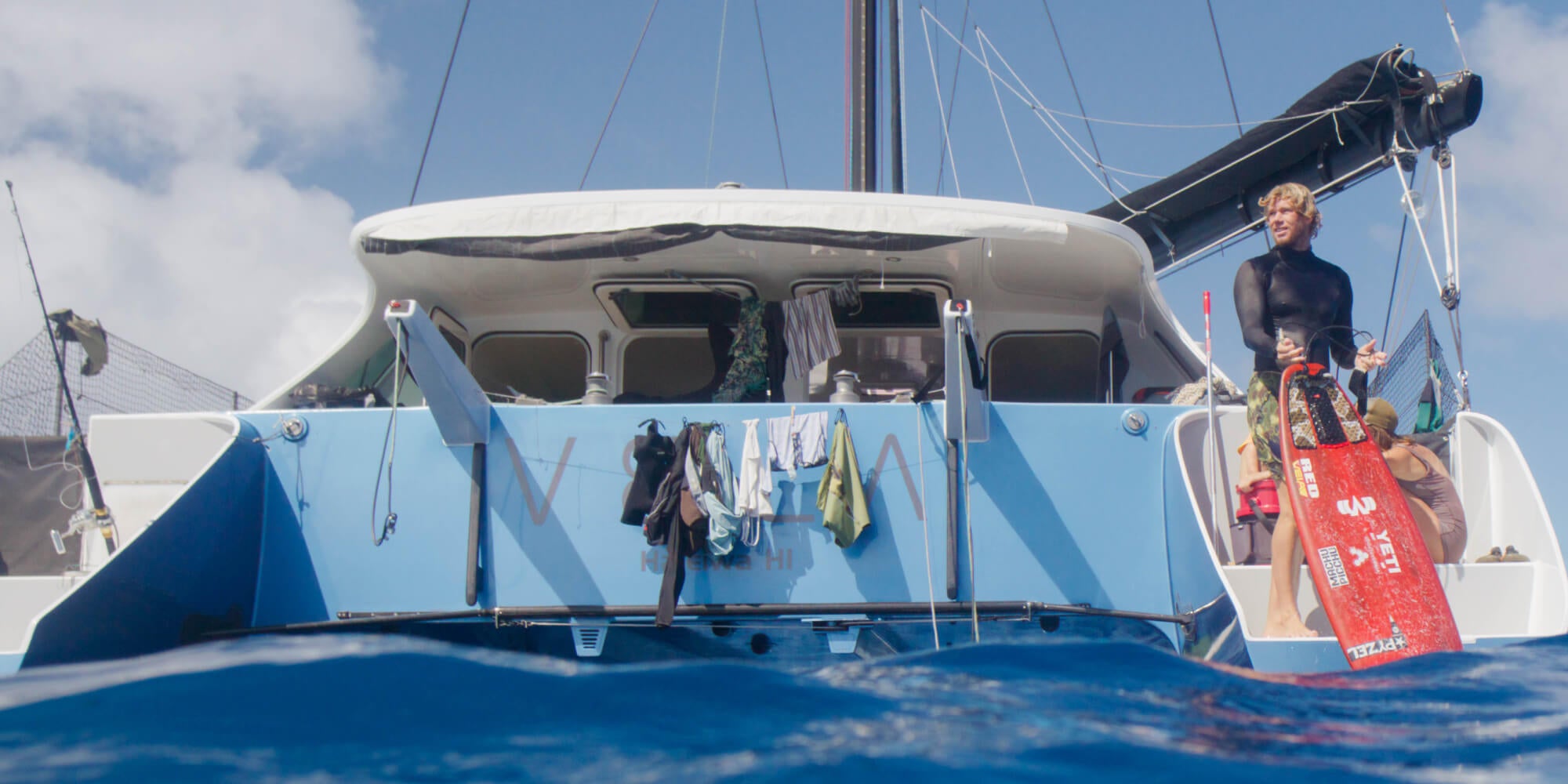 Person on a catamaran holding a surfboard  with a blue sky and water background