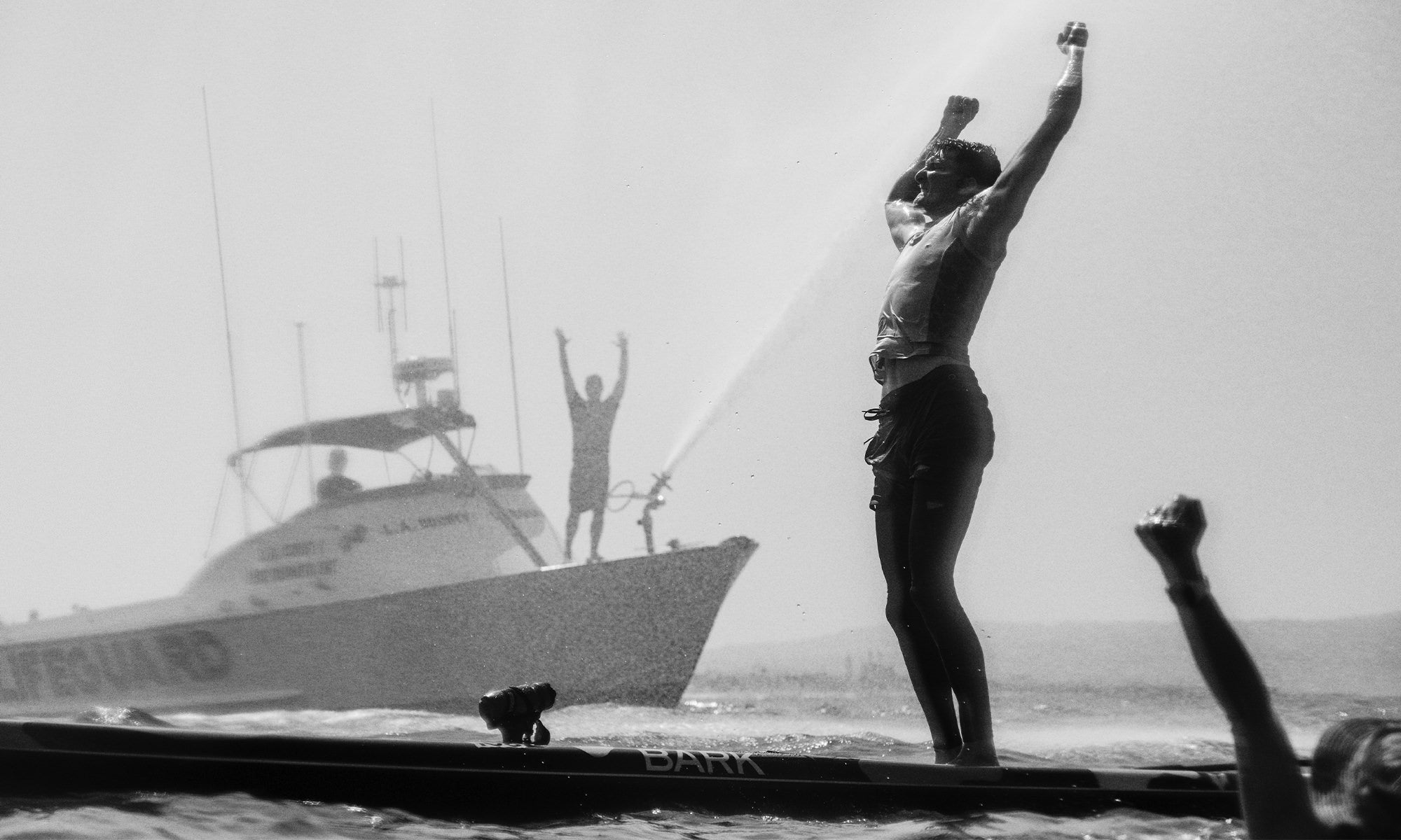 Black and white image of Jack Bark standing on his paddle board with his arms over his head claiming victory at the Catalina Classic Paddlbeoard race, with the lifeguard boat in the background spraying water and lifeguards cheering for Jack