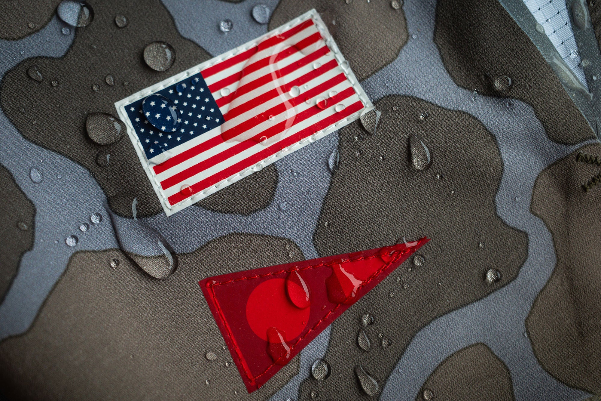 Zoomed in shot of camouflage board short fabric with two emblems, the top emblem is a small United States flag, the bottom is a red triangular burgee. The fabric has water droplets on it