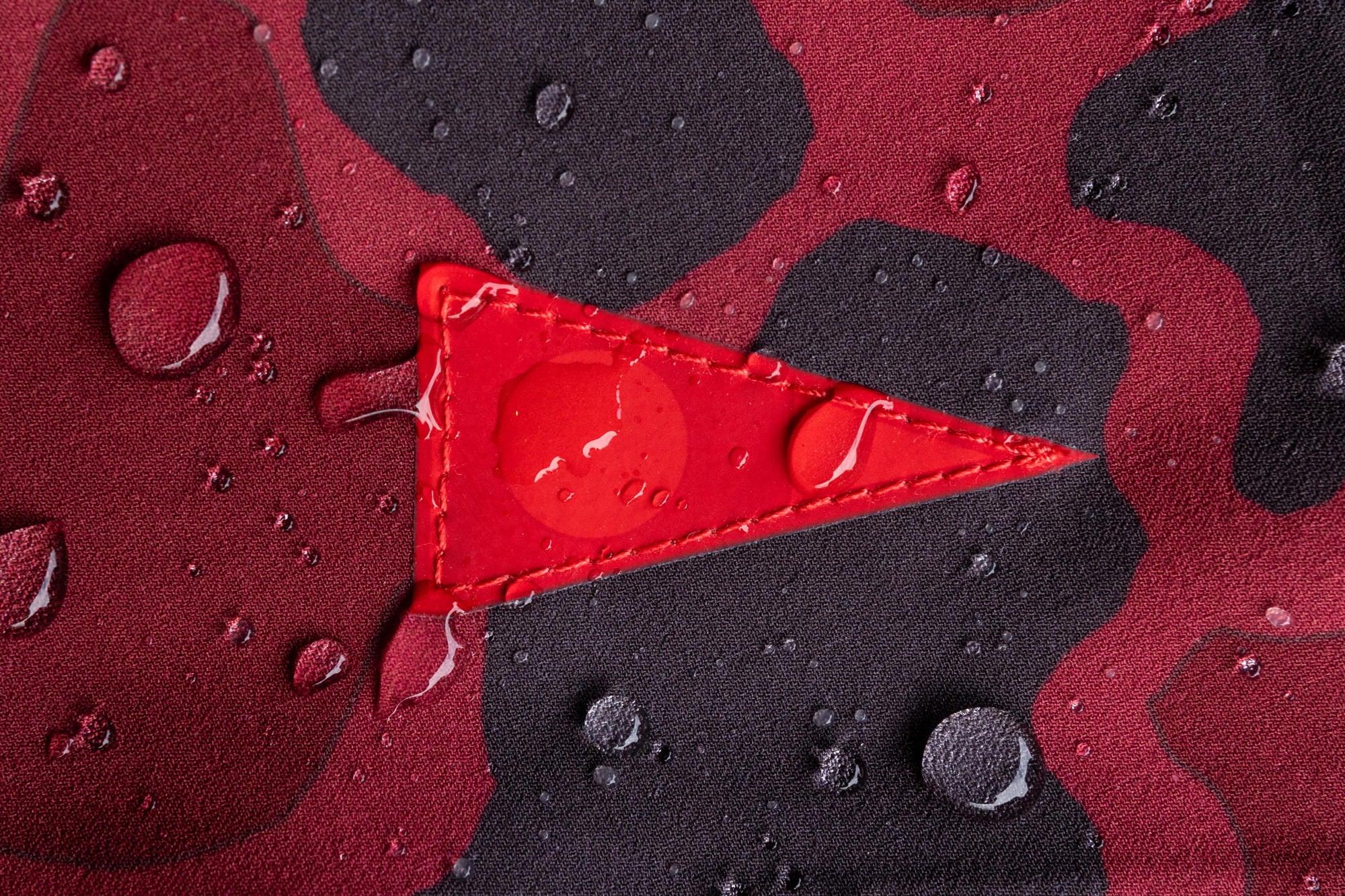 Zoomed in color image of maroon and black camo boardshort fabric, with the Florence Burgee-red triangular patch-and water droplets