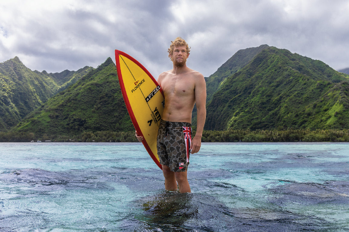 John John Florence standing on shallow reef in Tahiti with mountainous backdrop, wearing camouflage boardshorts with a Hawaiian flag side panel and holding a red and yellow surfboard