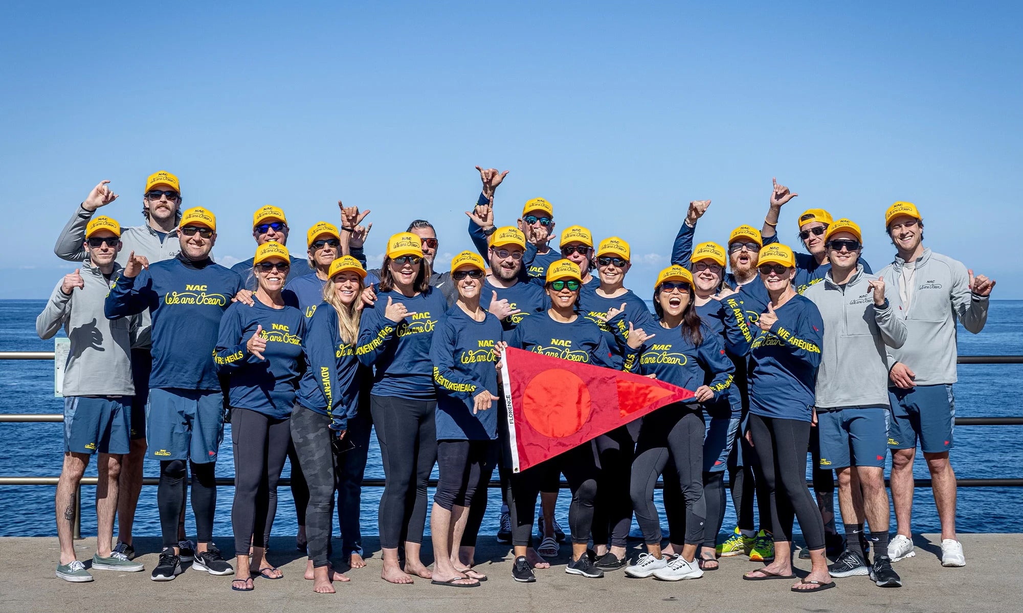 Group of people from We Are Ocean, wearing Florence x We Are Ocean Hats and shirts, standing and throwing shakas with the ocean in the background. Three people in the front center are holding a large triangular red Burgee flag