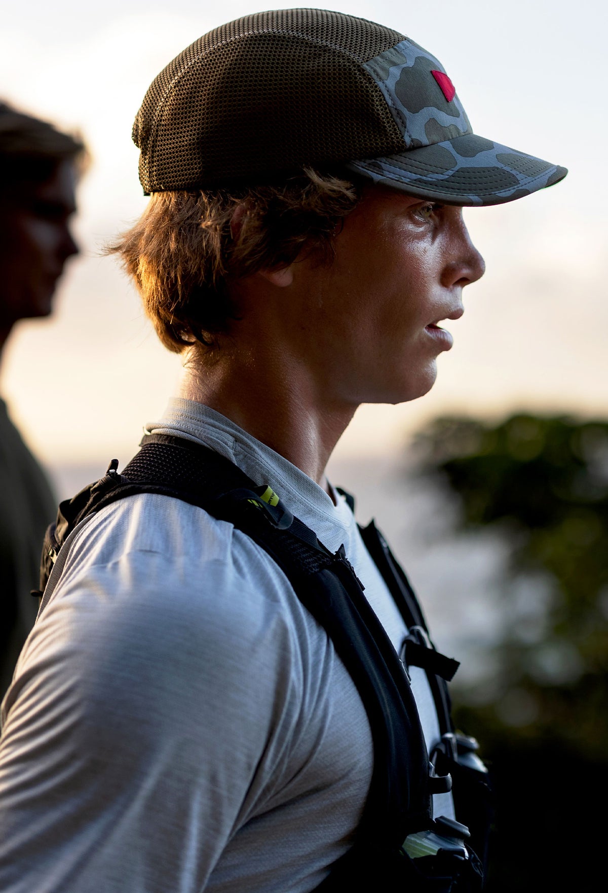 Side profile of a male athlete in the foreground, wearing a camouflage Florence Hat, who appears to have just finished a hike