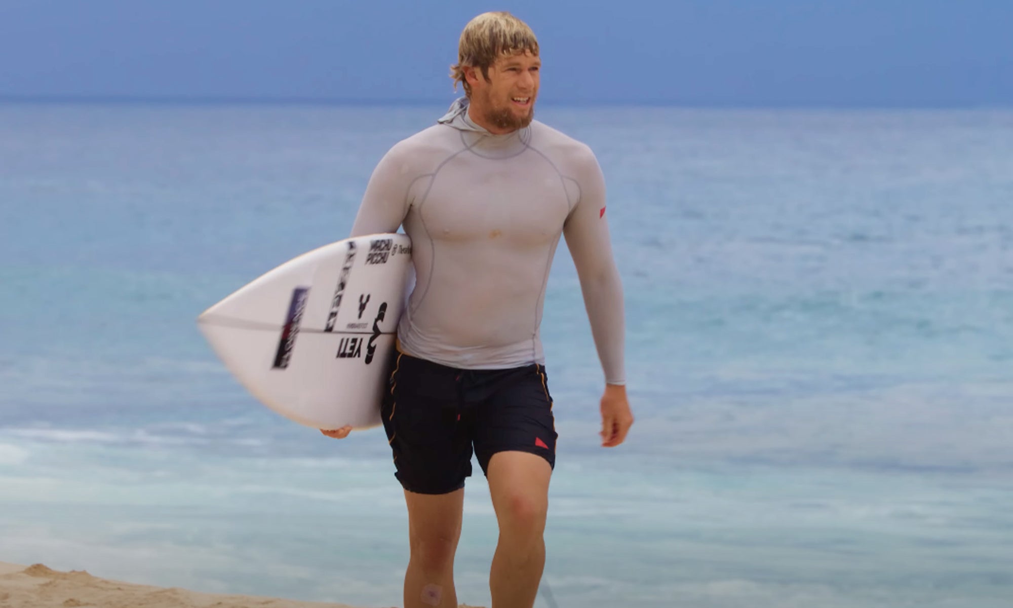 John Florence walking up the beach with a surfboard under his arm, wearing boardshorts and a rashguard