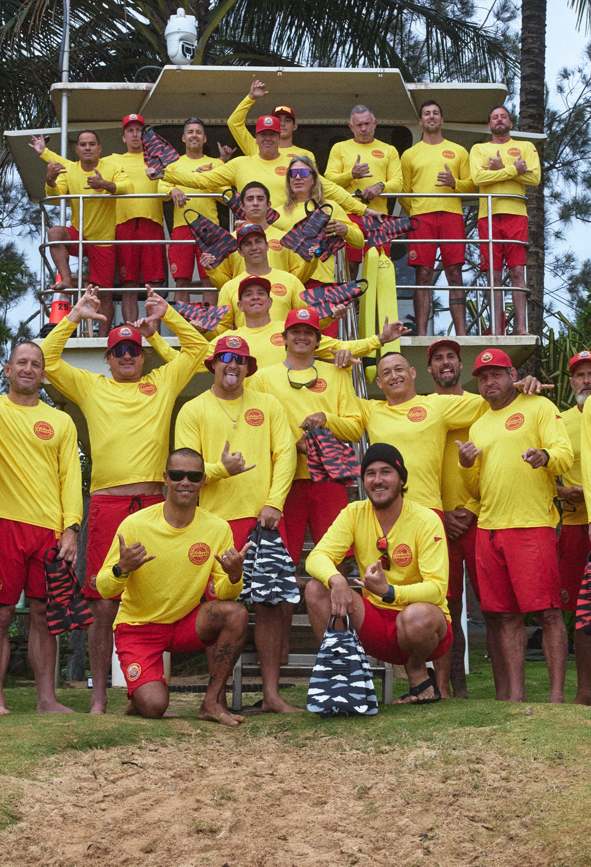 Image of the North Shore Lifeguards standing in front of a lifeguard tower, wearing yellow shirts, red boardshorts, red hats, holding their swim fins and throwing shakas