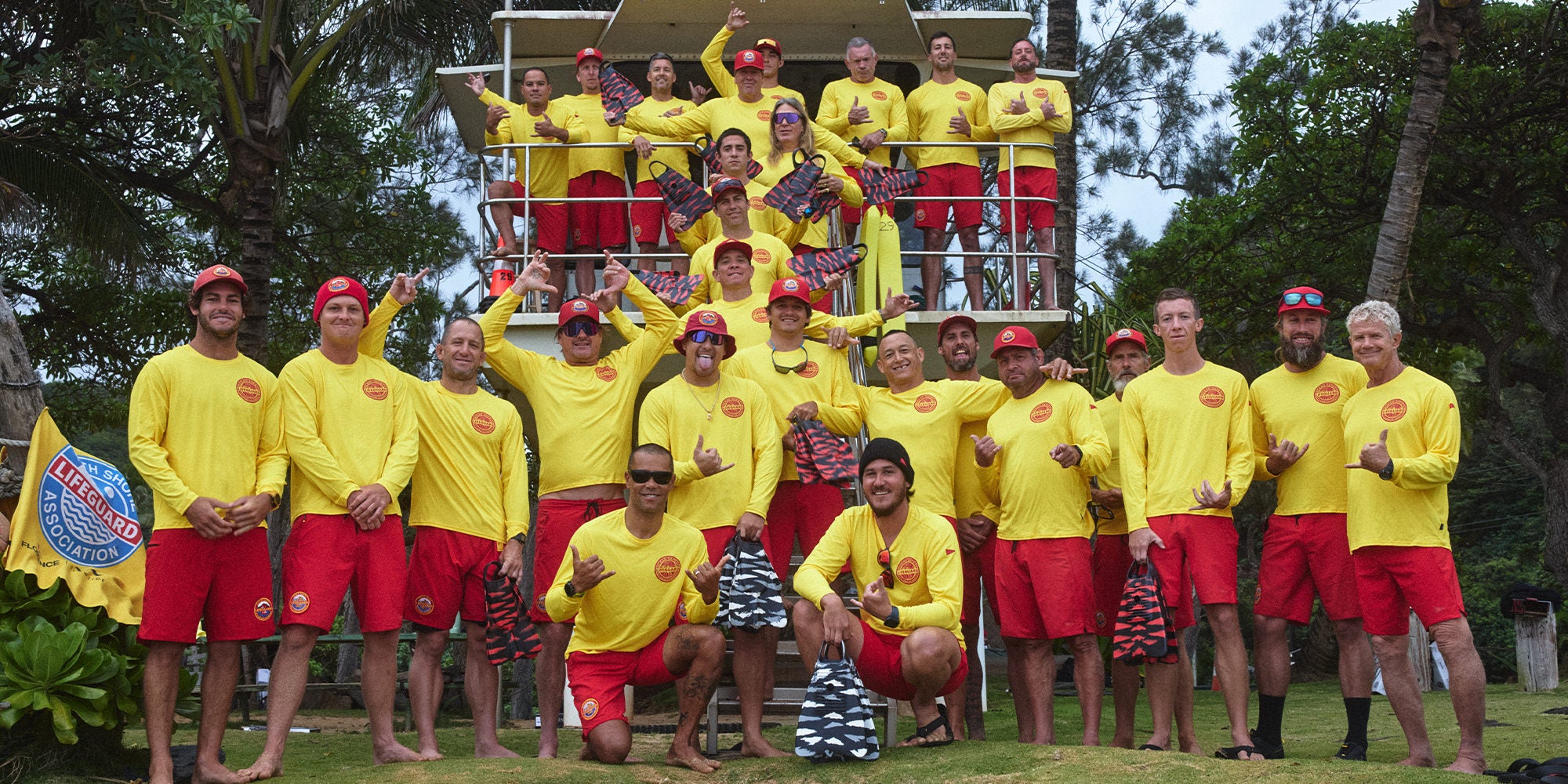 Image of the North Shore Lifeguards standing in front of a lifeguard tower, wearing yellow shirts, red boardshorts, red hats, holding their swim fins and throwing shakas
