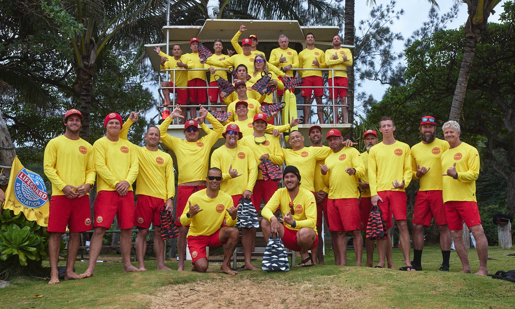 Image of the North Shore Lifeguards standing in front of a lifeguard tower, wearing yellow shirts, red boardshorts, red hats, holding their swim fins and throwing shakas