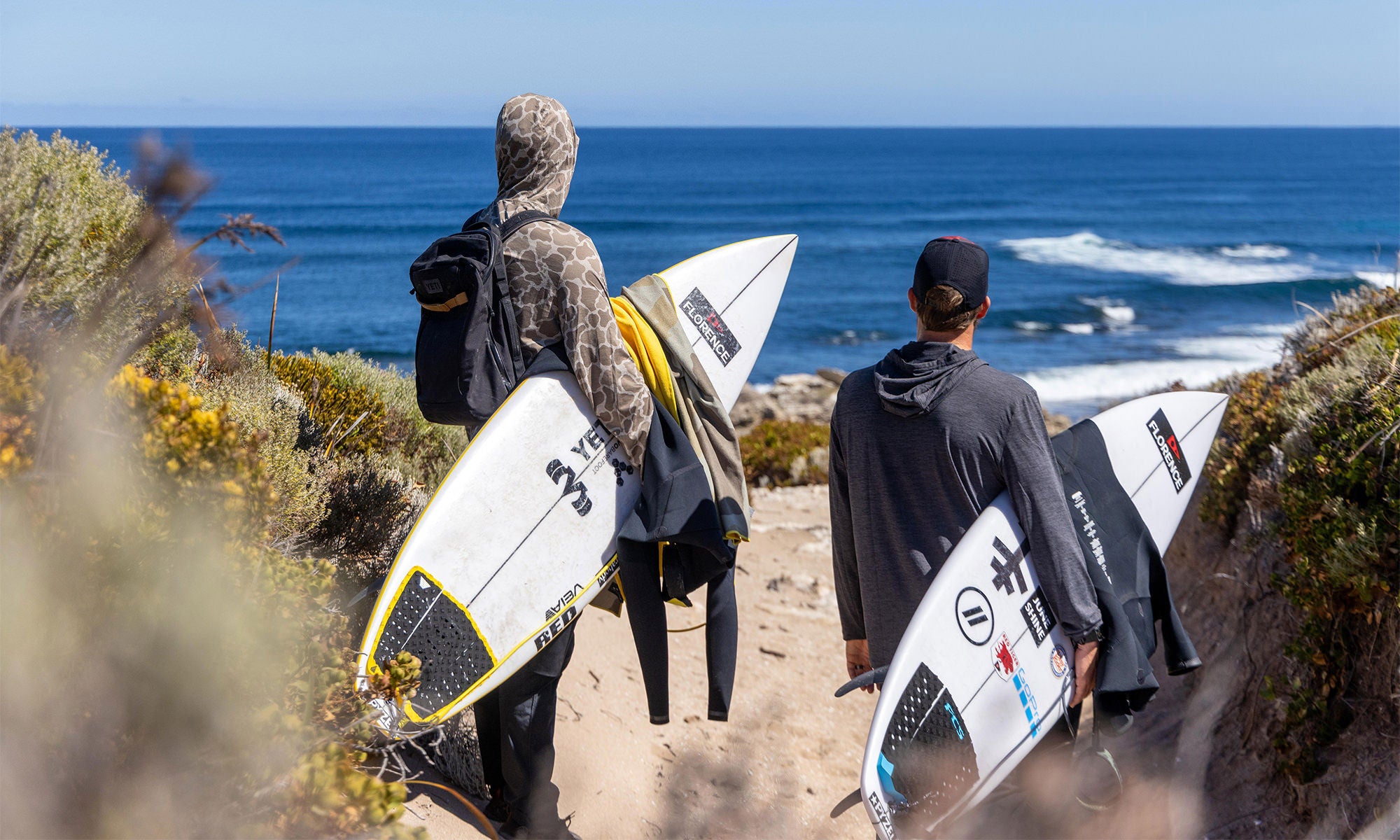 Two people holding surfboards, on the sand with brush on the sides of them, looking towards the ocean in the horizon