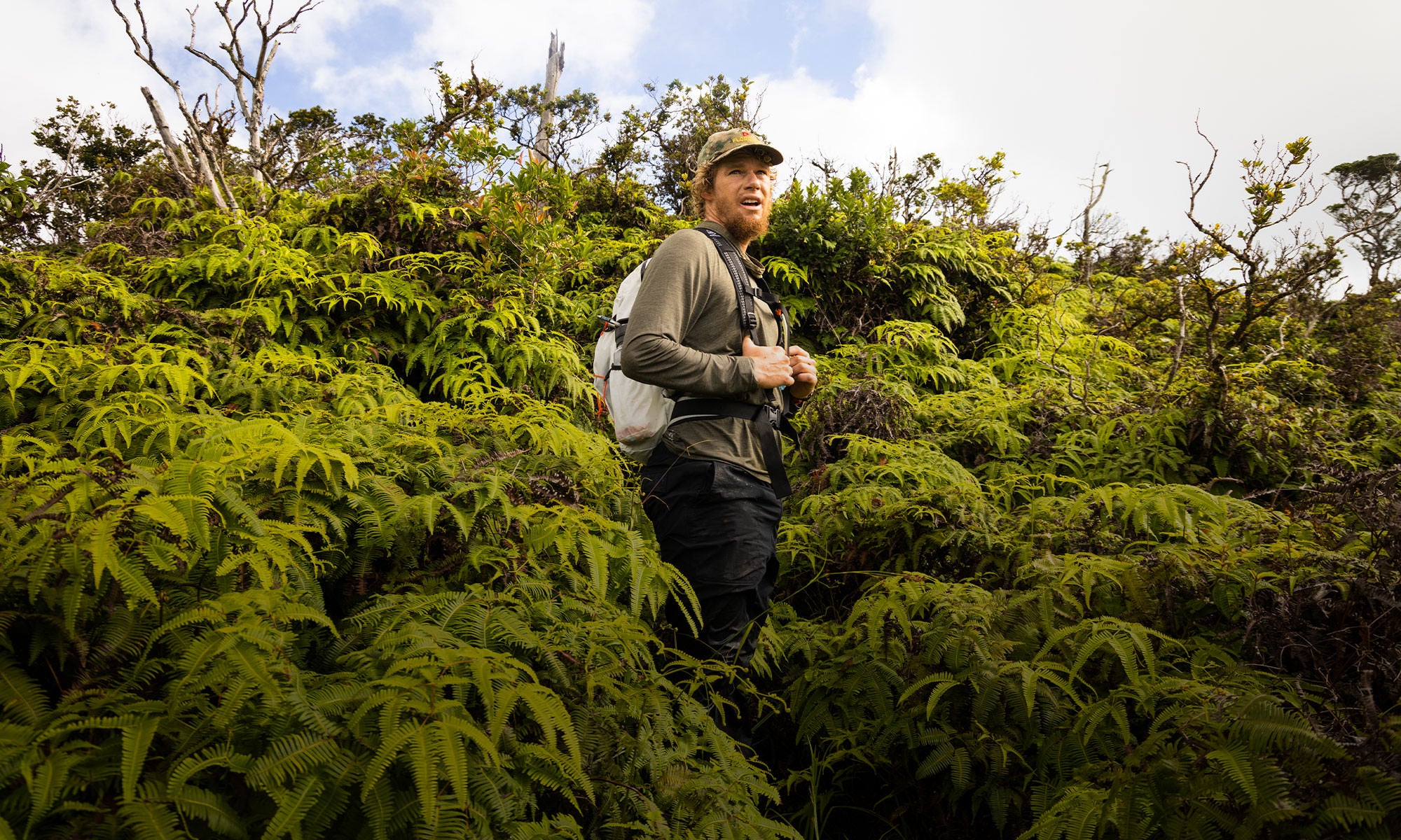 John John Florence hiking through dense Hawaiian foliage, wearing a green long sleeve shirt, black pants, a camouflage Florence hat, and a backpack