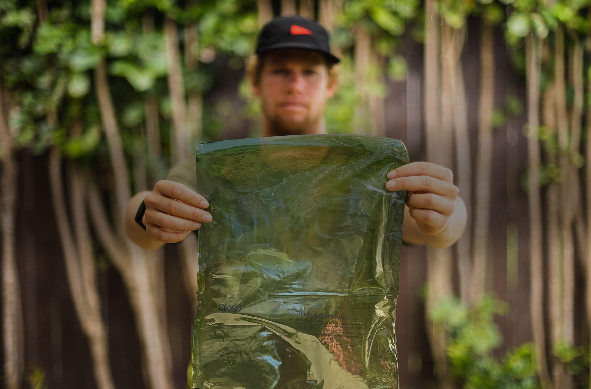 Man wearing black hat holding up a bag made from recycled seaweed, with a wooded tree background. The green bag is in focus, while the man is out of focus